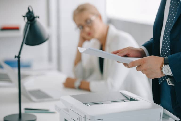 Selective focus of businessman holding papers above printer, woman in background at desk